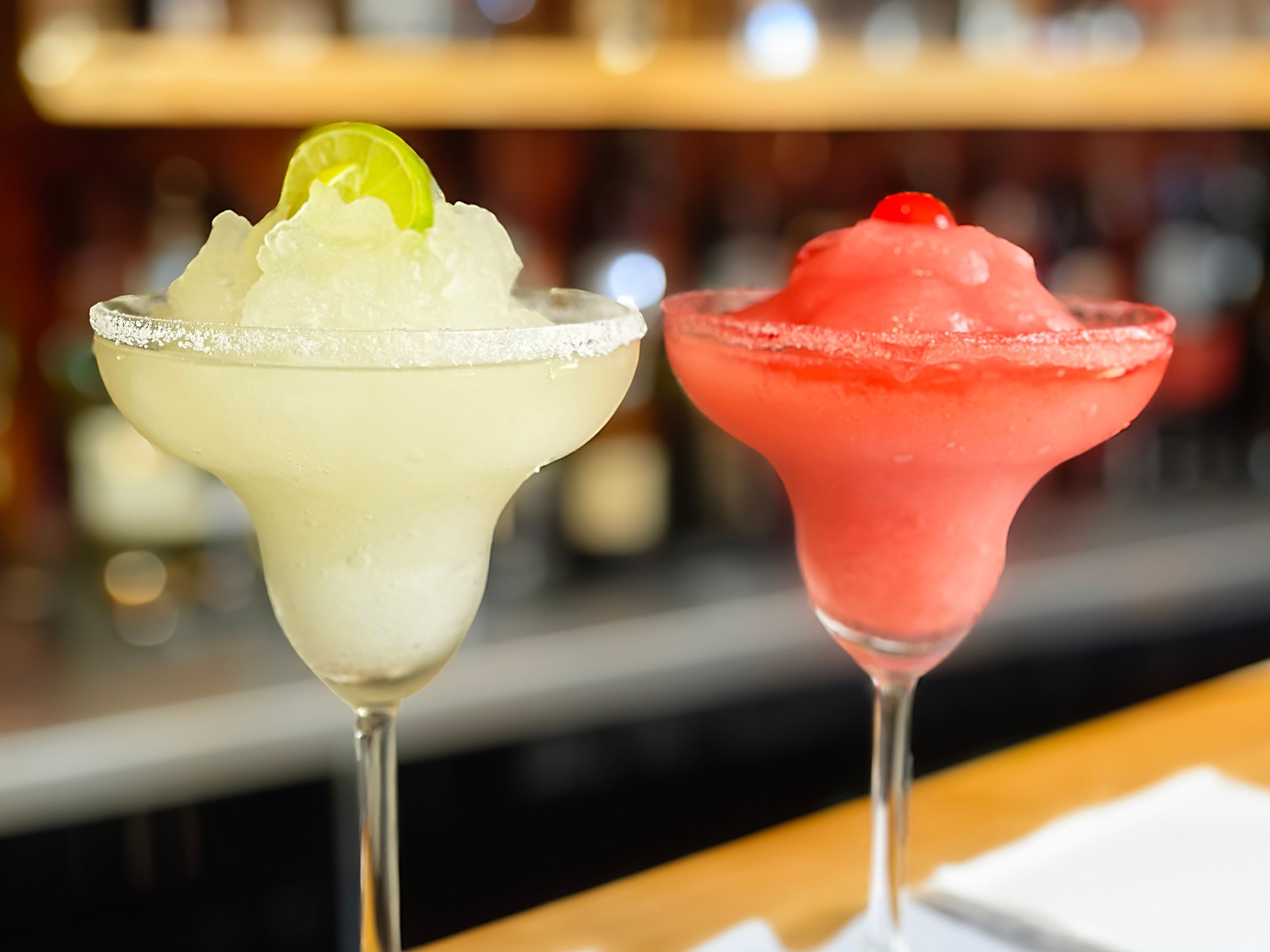 Close-up landscape image of frozen cocktails at bar,  lime and strawberry iced margaritas against blurred bar background, margarita glass with salted rim and fruit garnishes.